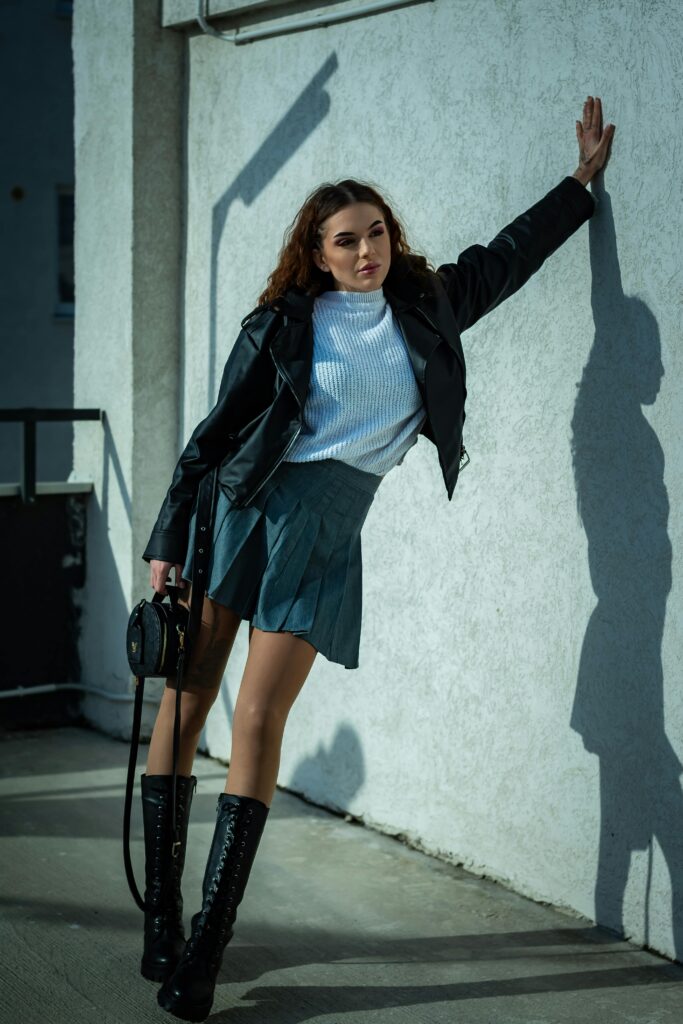 Stylish woman in leather jacket and skirt leaning against a sunlit wall outdoors.